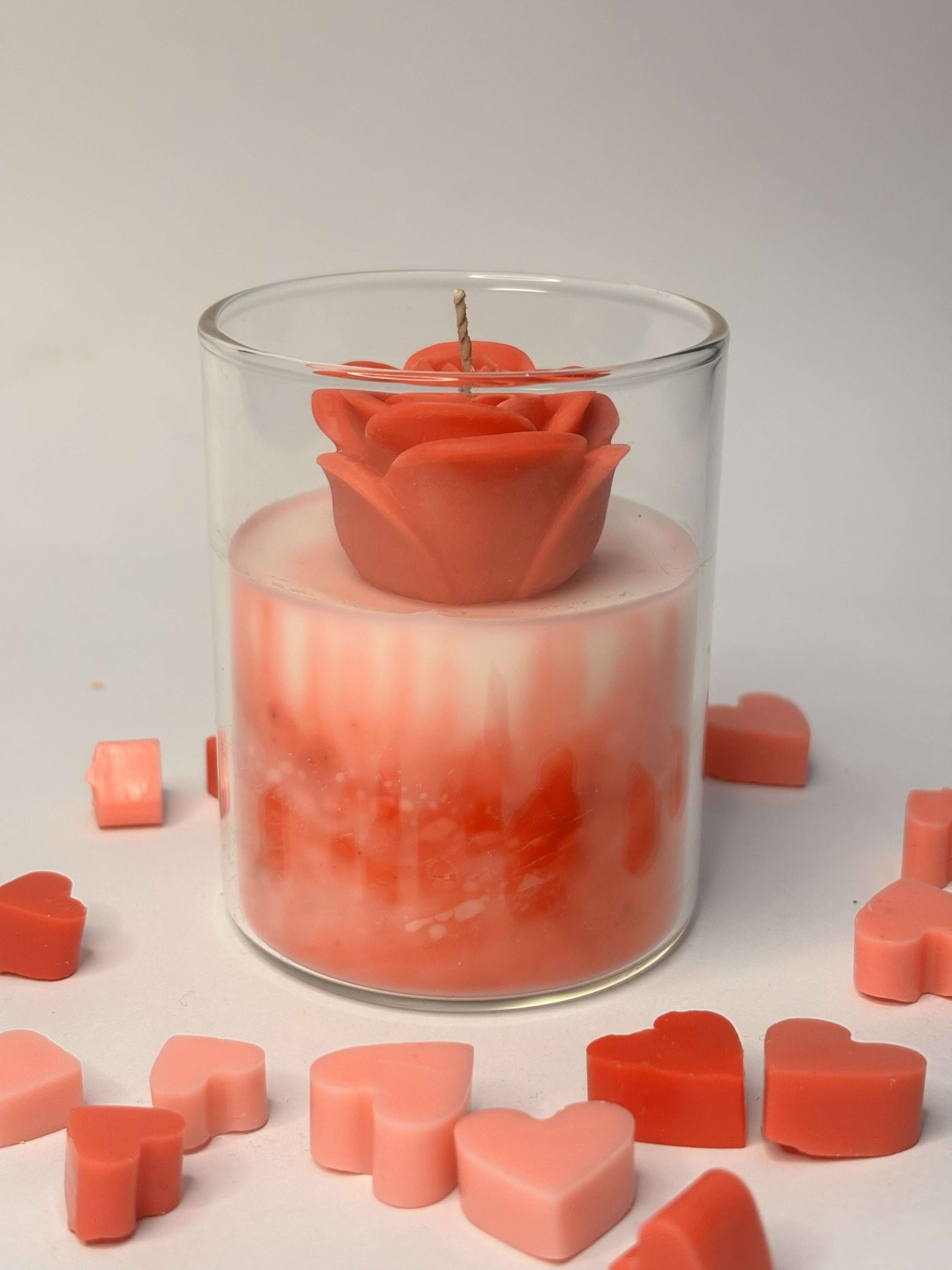 Handcrafted layered beeswax candle in a clear borosilicate jar, featuring a marbled coral and ivory wax body topped with a single sculpted rose-shaped bloom and surrounded by small red and pink heart-shaped wax accents.