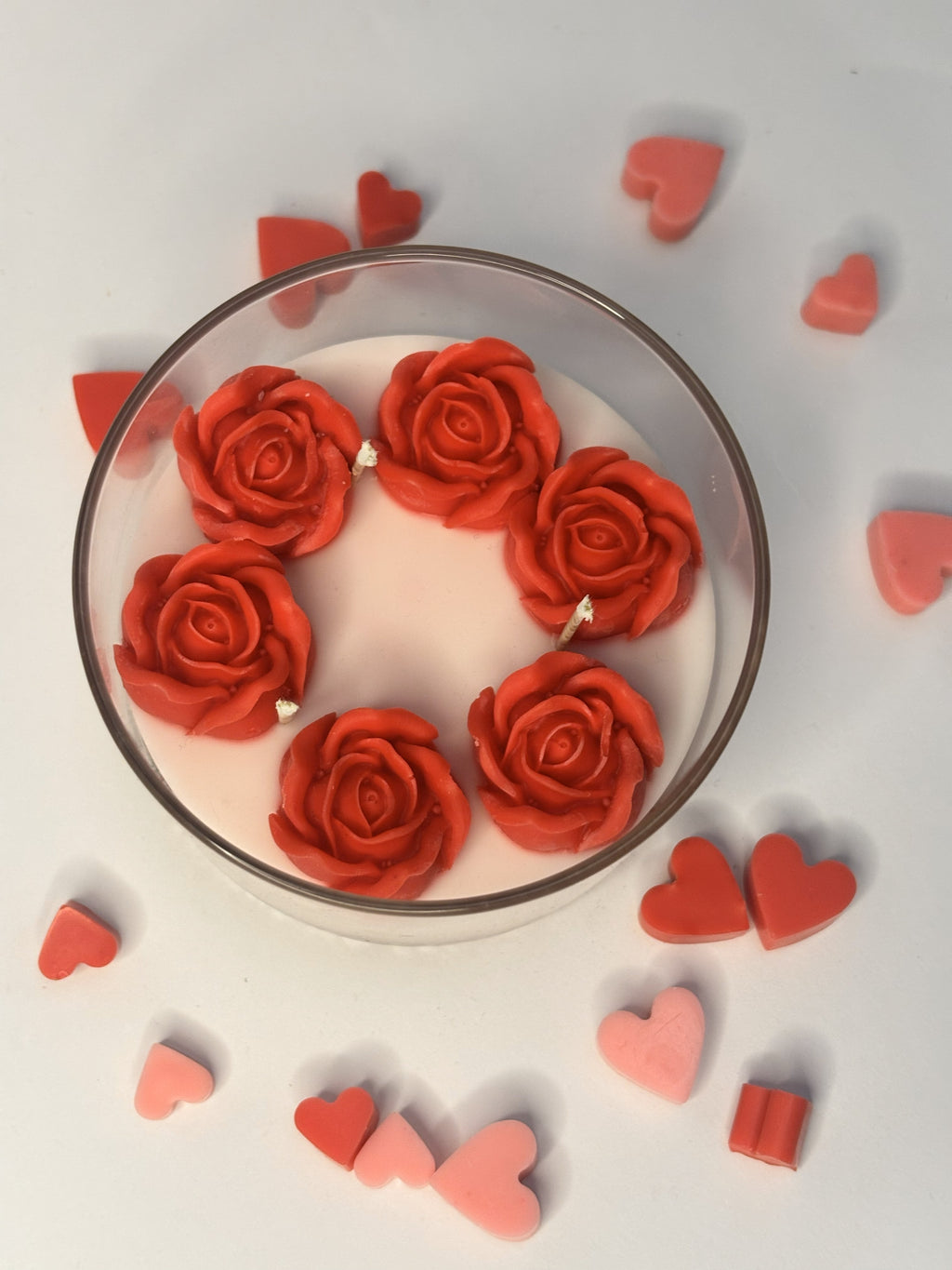 Top view of a layered beeswax candle in a clear borosilicate jar, featuring a smooth ivory wax base topped with six sculpted crimson rose-shaped blooms arranged in a circle, surrounded by small red and pink heart-shaped wax accents.