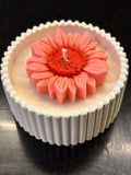 A close-up of a handcrafted beeswax candle in a white ribbed jar, topped with a sculpted pink sunflower-shaped beeswax flower and a centered cotton wick, set against a dark background.