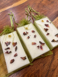 Wickory handcrafted beeswax wax sachets with pressed botanical petal decorations, individually scented and wrapped in sheer olive organza pouches, displayed on a wooden surface