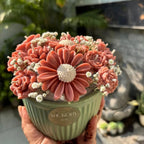 A hand holding the Wickory Blossom Posy: coral-pink sculpted beeswax flower candles with white filler flowers in a ribbed sage green pot with a gold Wickory label