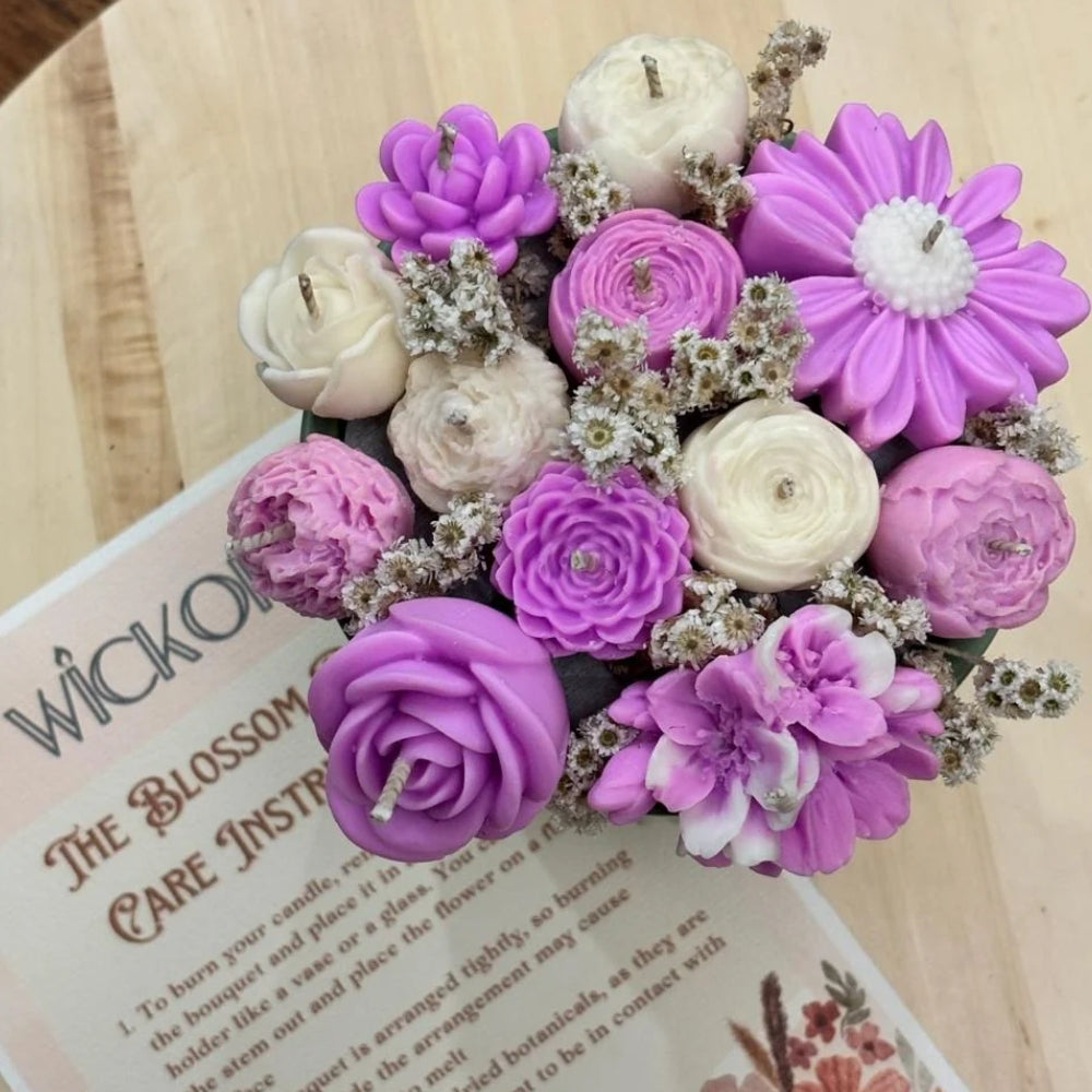 A bouquet-style arrangement of flower-shaped candles in purple, lavender, cream, and pink tones with small white filler flowers, photographed on a wooden surface over a printed candle care card.