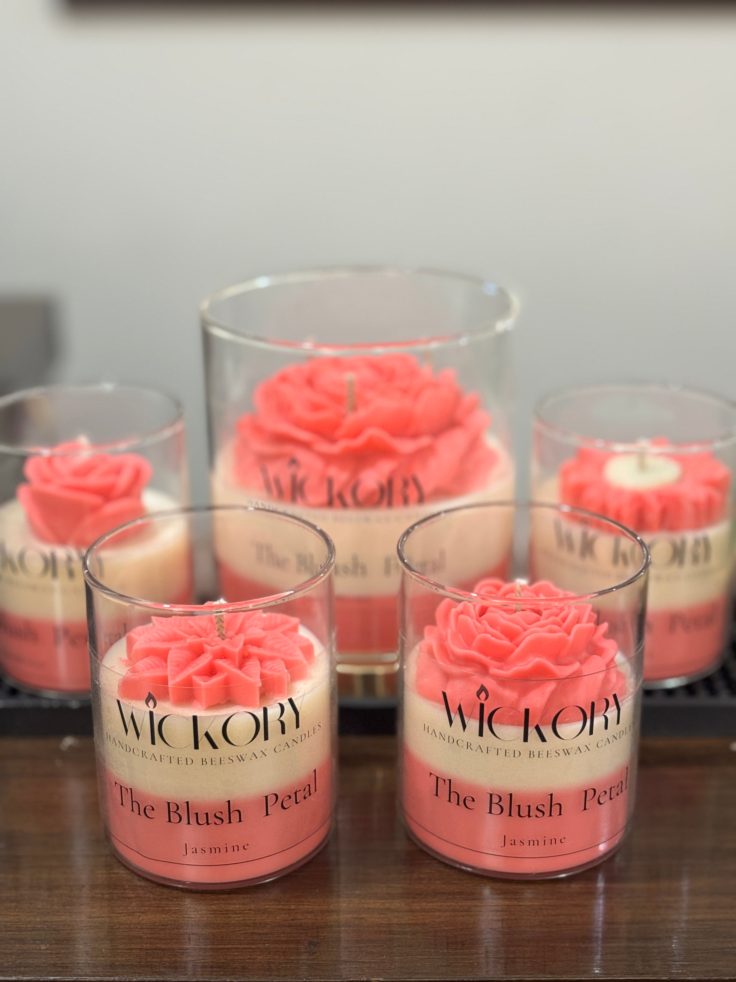 Five Wickory beeswax candles arranged on a wooden surface, featuring four small jars and one larger jar in the center, each with blush and ivory layered wax and sculpted floral tops in clear glass containers.