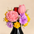 Cork & Bloom beeswax flower candle topper on a black wine bottle: pink rose, pink tulip, lilac and yellow blooms with baby's breath, on a beige background
