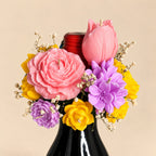 Cork & Bloom beeswax flower candle topper on a black wine bottle: pink rose, pink tulip, lilac and yellow blooms with baby's breath, on a beige background