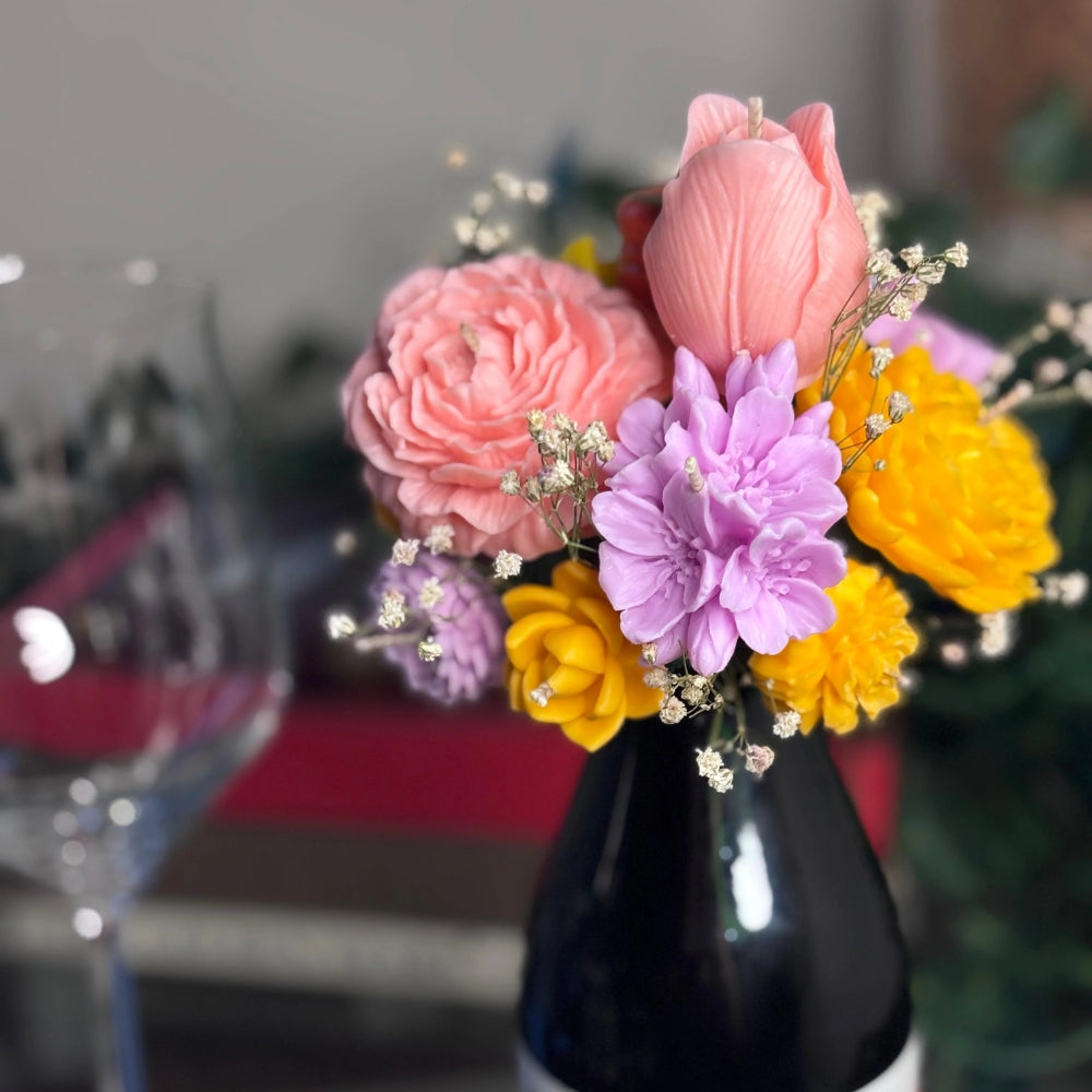 Cork & Bloom beeswax flower topper with pink rose, pink tulip, lilac and yellow blooms on a black wine bottle, beside a wine glass