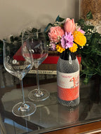 Cork & Bloom beeswax flower candle topper on a netted wine bottle, with pink rose, pink tulip, lilac and yellow blooms, styled beside two wine glasses