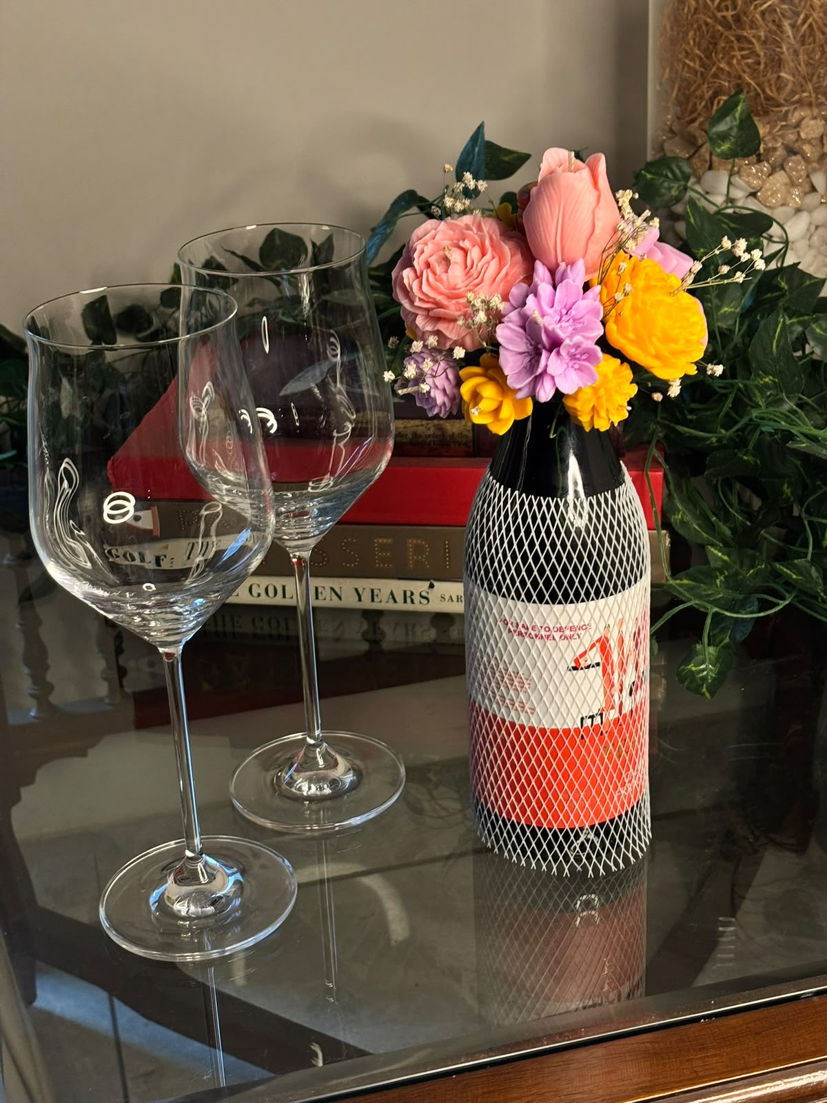 Cork & Bloom beeswax flower candle topper on a netted wine bottle, with pink rose, pink tulip, lilac and yellow blooms, styled beside two wine glasses
