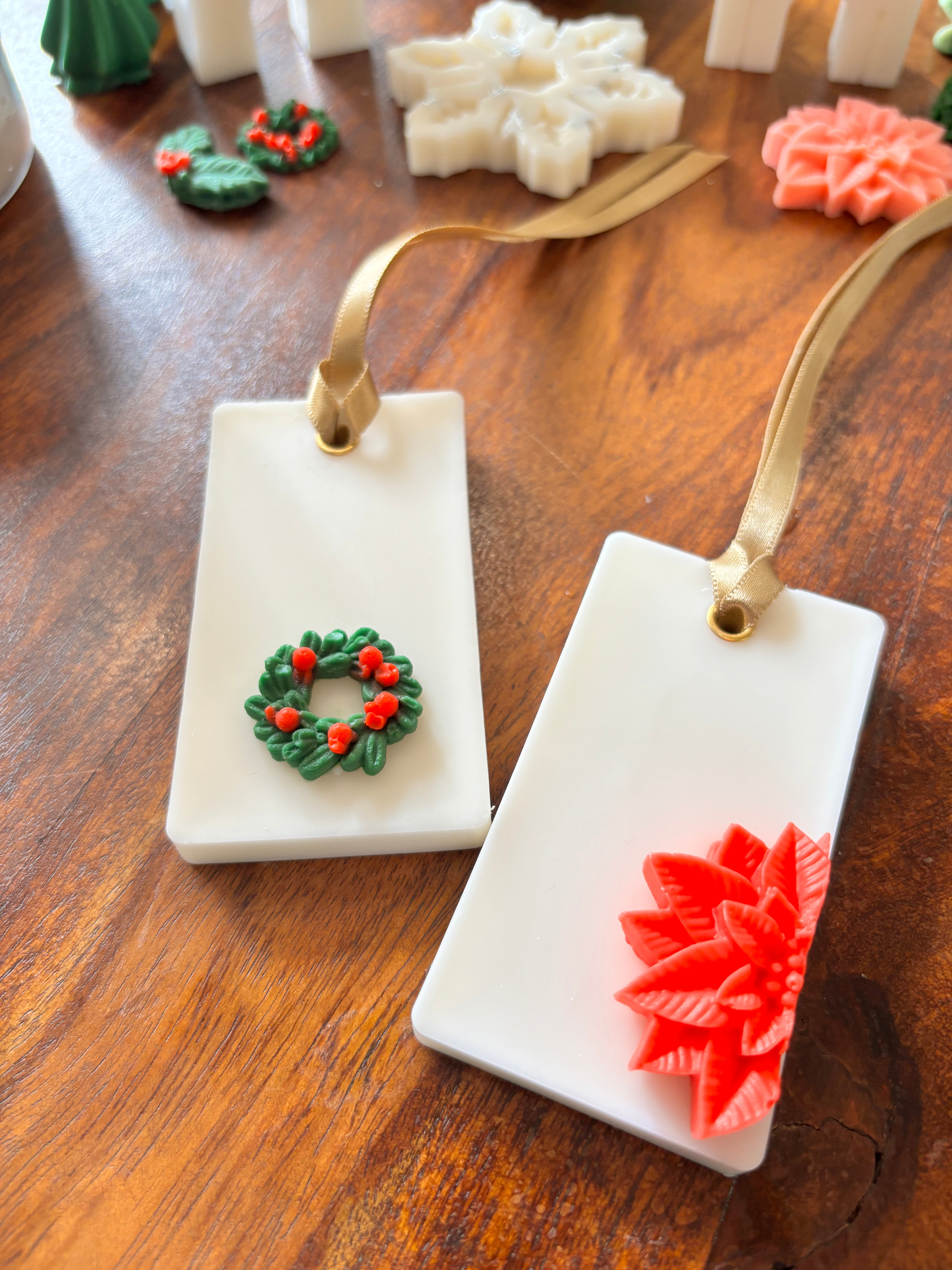 2 Handcrafted rectangular beeswax sachets with ribbon loops, featuring festive wax motifs including a green wreath and a red poinsettia, displayed on a wooden surface.