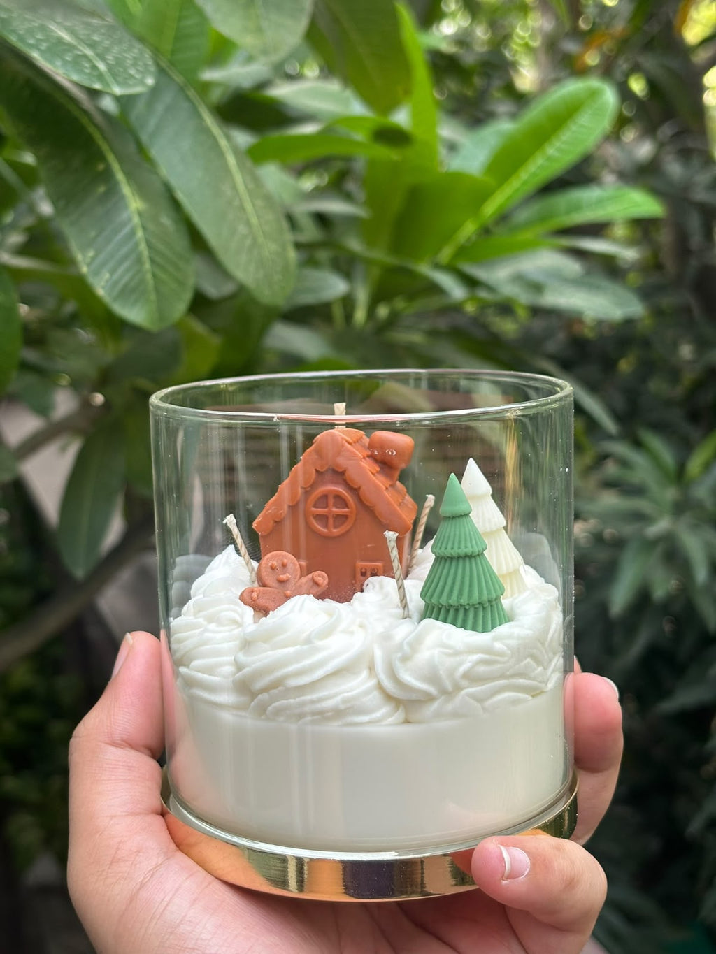 The Frosted Wonderland beeswax Christmas candle held in hand: sculpted gingerbread house, gingerbread figure and green and white trees on whipped white wax in a gold-based glass jar
