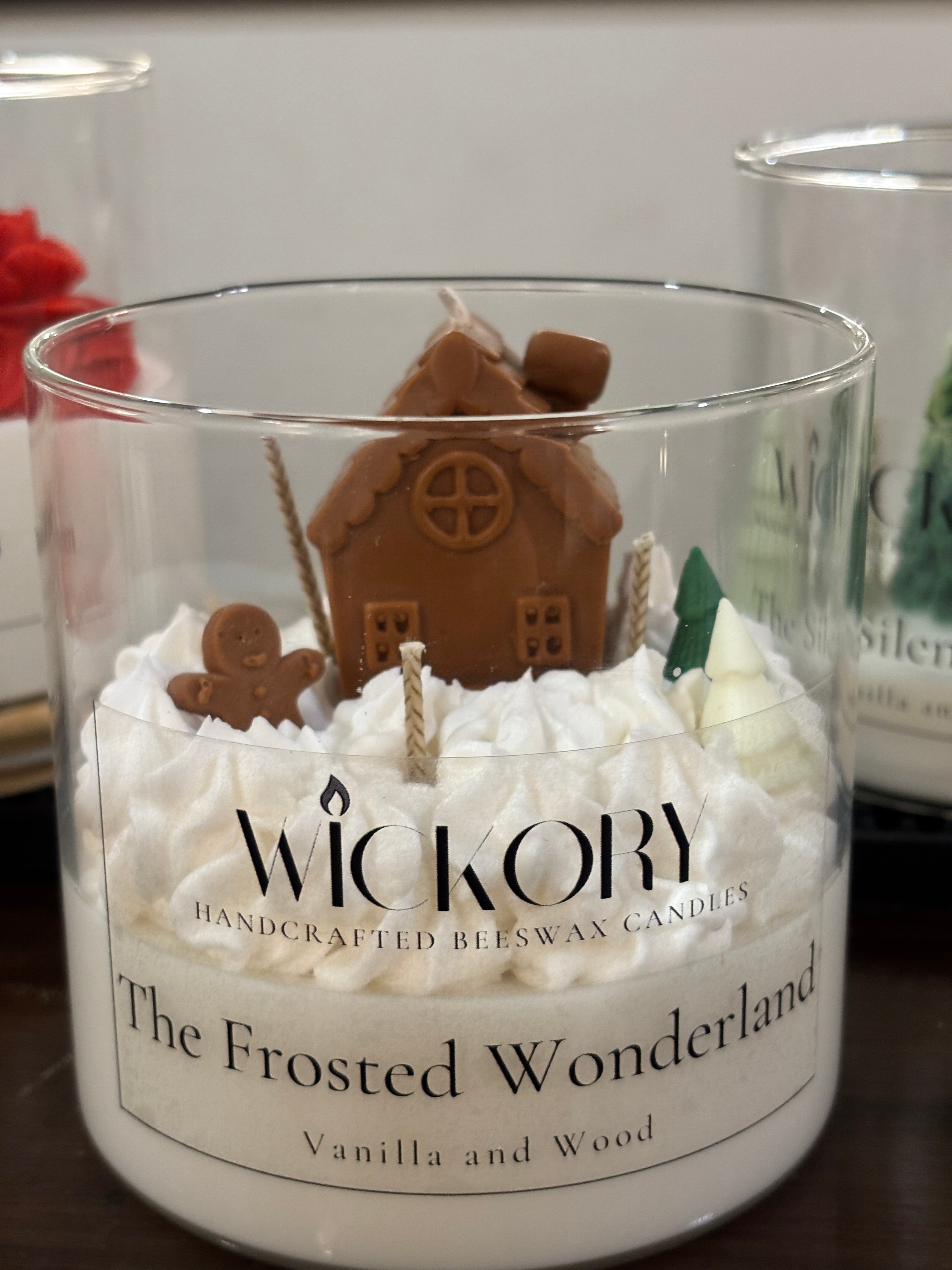 Handcrafted Wickory beeswax candle in a clear glass jar, featuring whipped white wax topped with a sculpted gingerbread house, a small gingerbread figure, and miniature tree details.