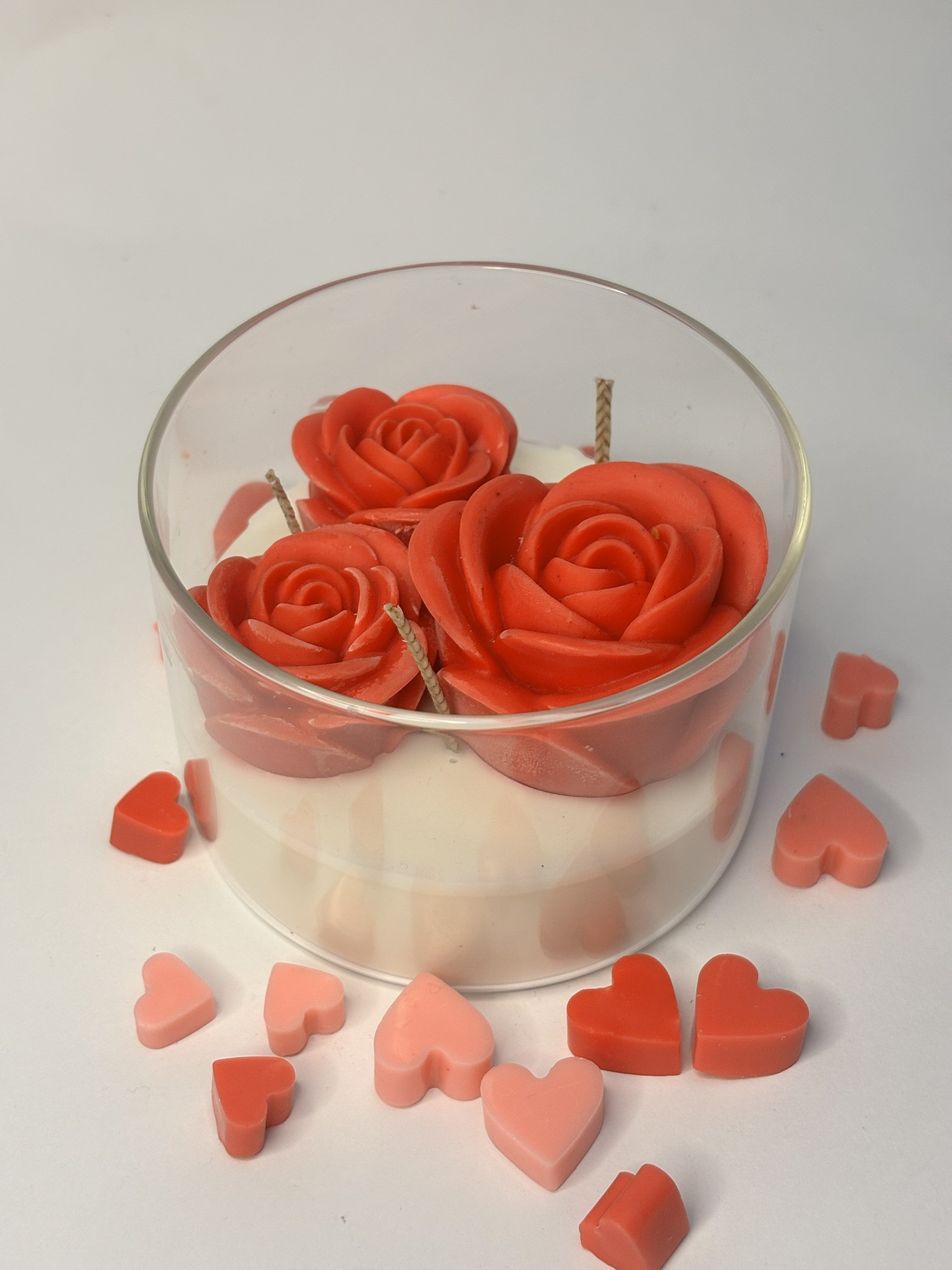 Handcrafted beeswax candle in a clear borosilicate jar, featuring three sculpted red rose-shaped blooms set on a smooth ivory wax base, surrounded by small heart-shaped wax accents.