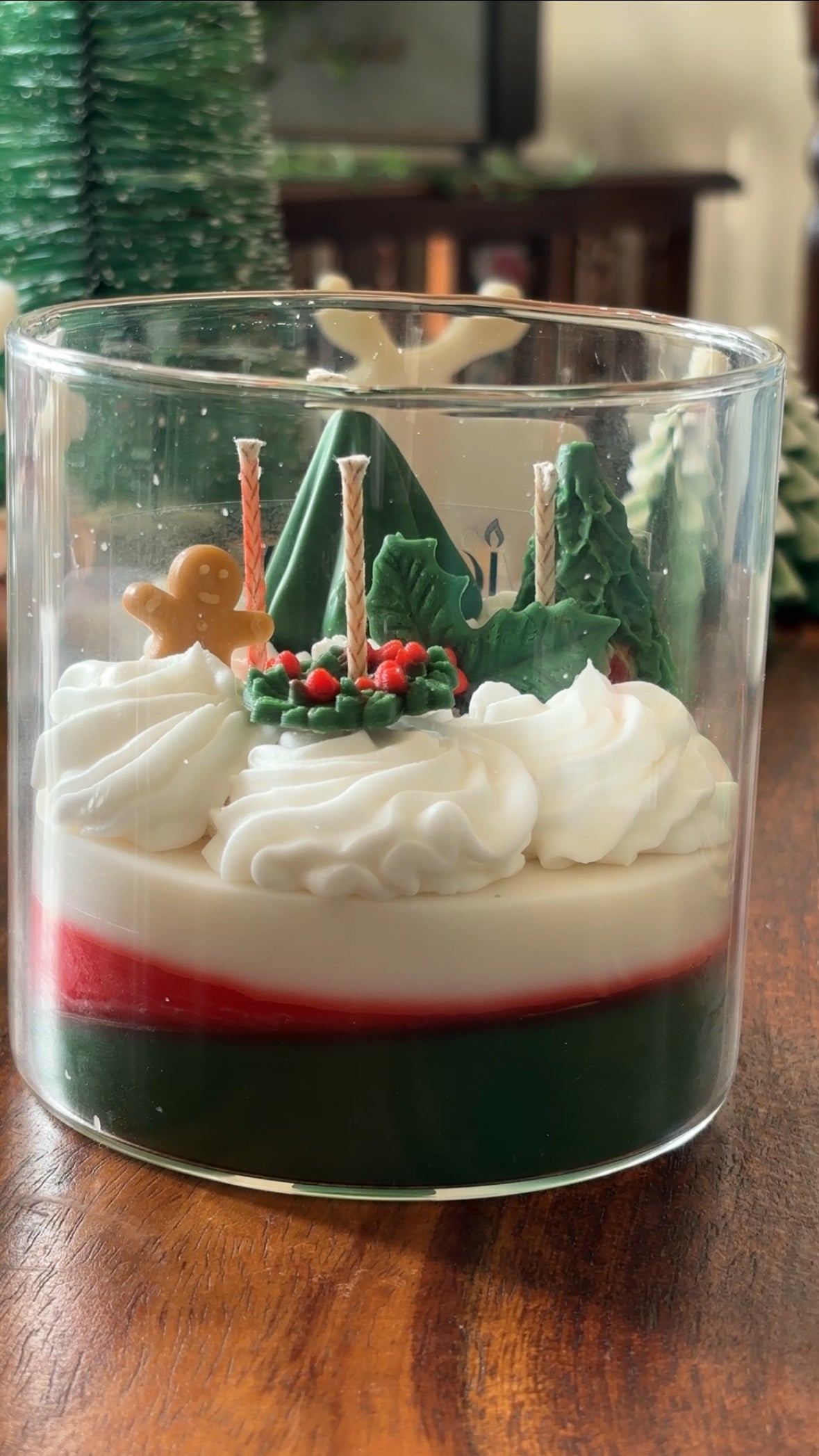 Handcrafted layered beeswax candle in a clear glass jar featuring green and red wax layers topped with textured white wax swirls and festive sculpted details including a gingerbread figure, holly leaves, berries, and tree shapes.