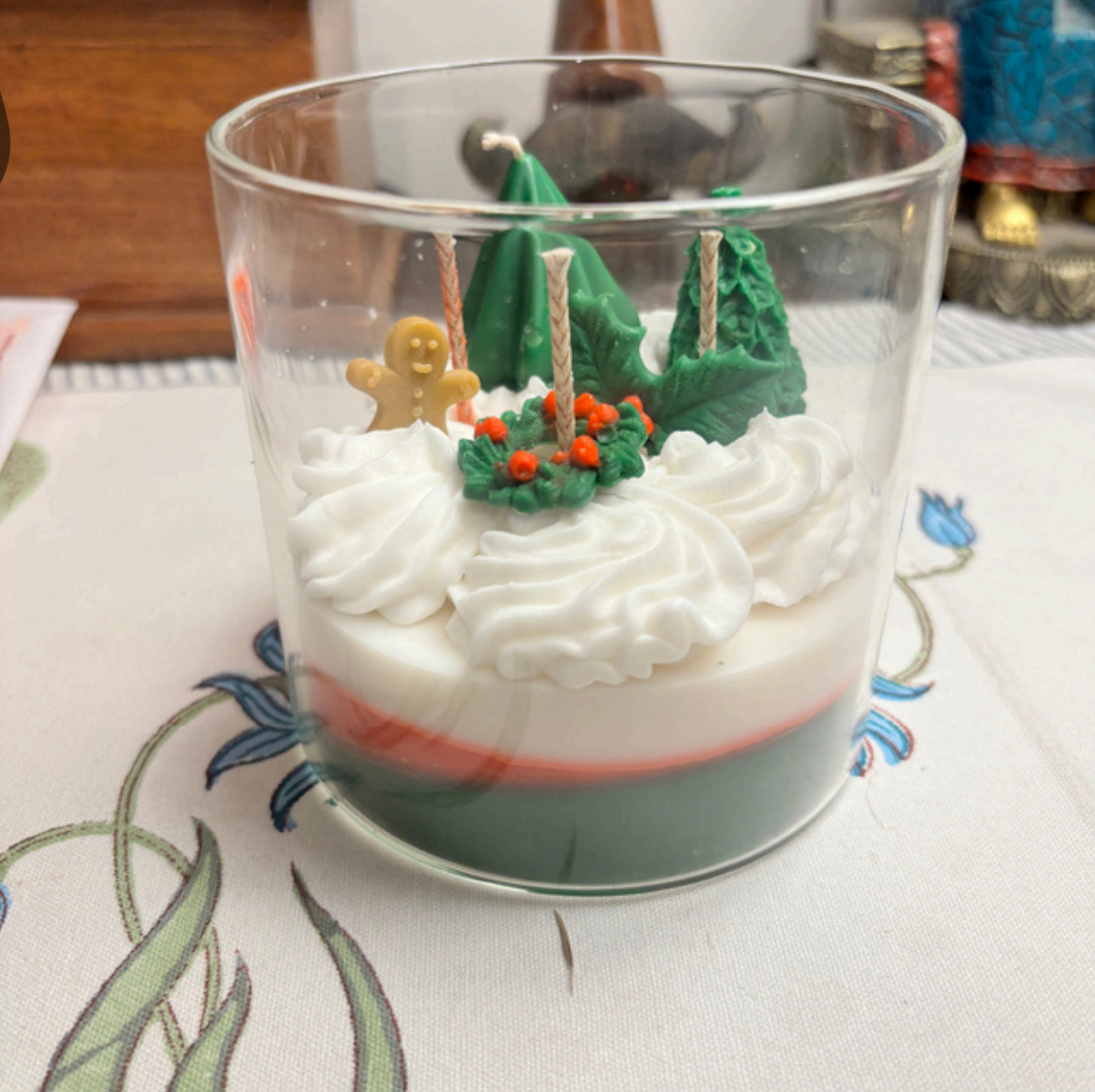 Handcrafted layered beeswax candle in a clear glass jar featuring green and red wax layers topped with textured white wax swirls and festive sculpted details including a gingerbread figure, holly leaves, berries, and tree shapes.