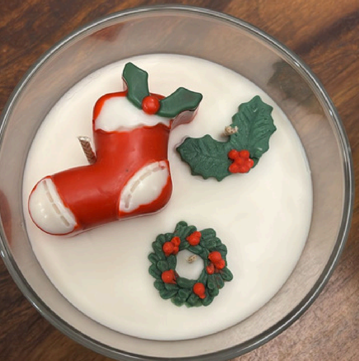 Top view of a handcrafted beeswax candle in a clear glass jar with an ivory wax base, decorated with festive sculpted wax details including a red stocking, holly leaves, and a green wreath.