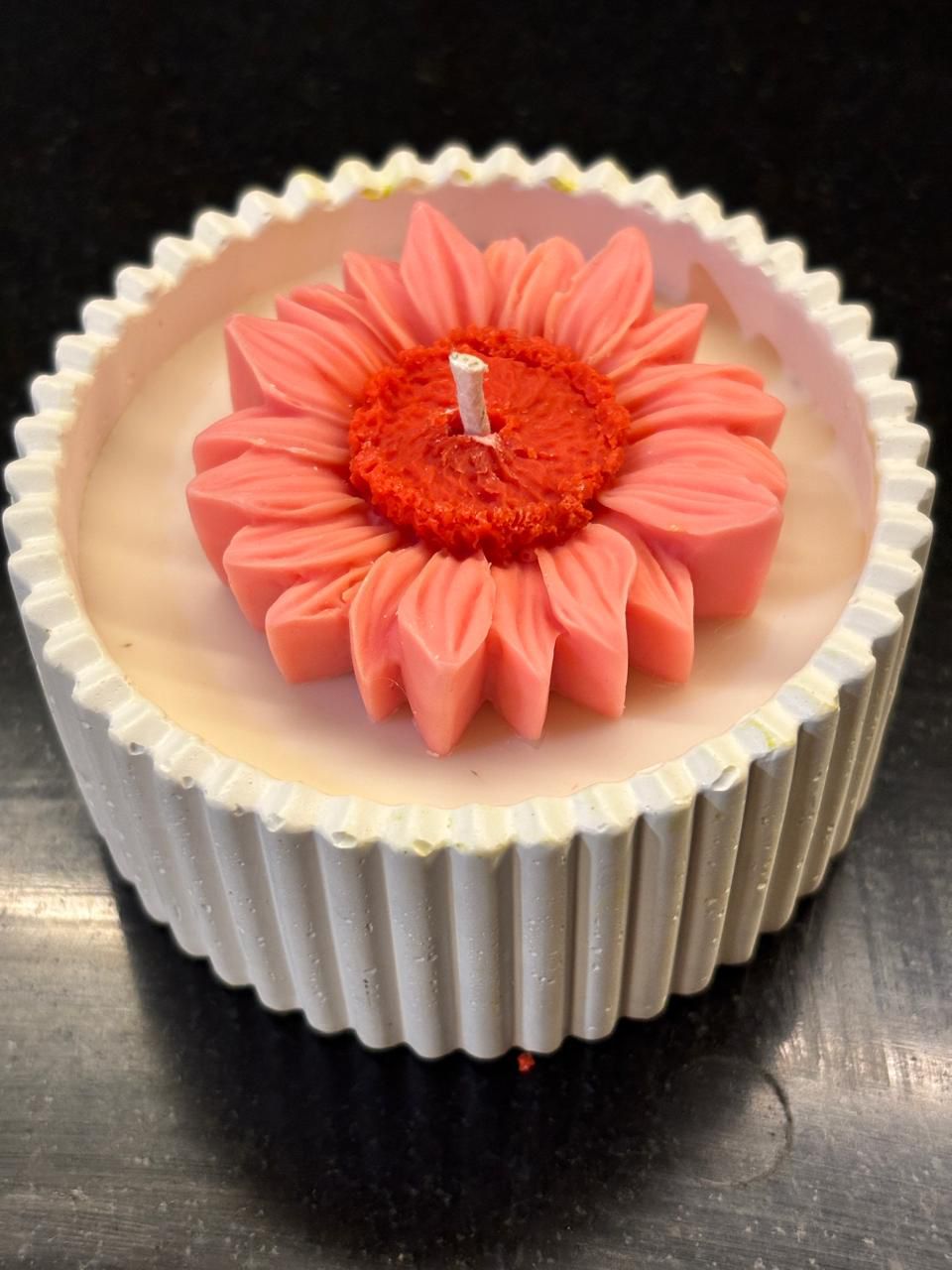 A close-up of a handcrafted beeswax candle in a white ribbed jar, topped with a sculpted pink sunflower-shaped beeswax flower and a centered cotton wick, set against a dark background.